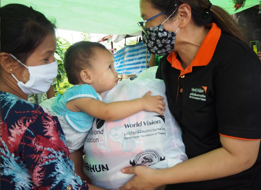 Un miembro del personal usa una mascarilla para entregar una bolsa para apoyar a las familias y prevenir la propagación de la COVID-19 en Tailandia. La mujer que recibe la bolsa sonríe mientras su hijo se aferra a la bolsa.