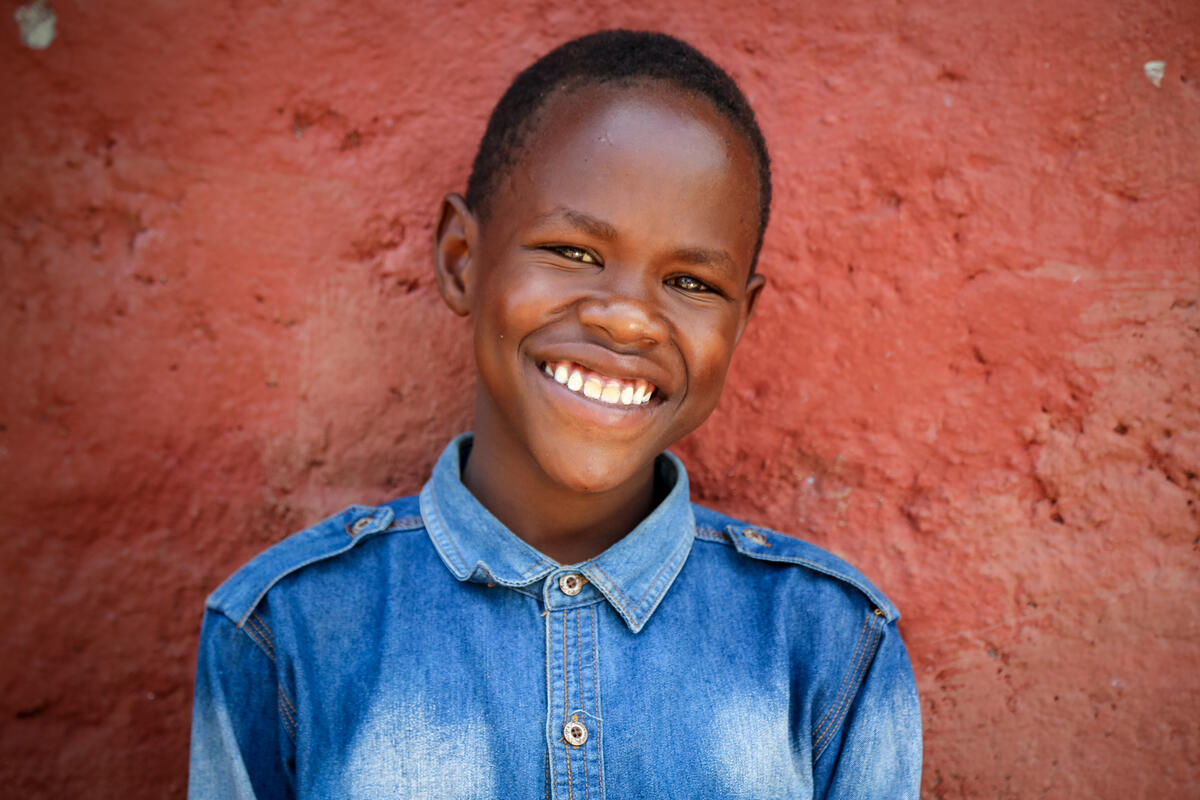 Niño apadrinado de 12 años en Kenia sonríe a la cámara, vestido con una camiseta vaqueras frente a una pared roja