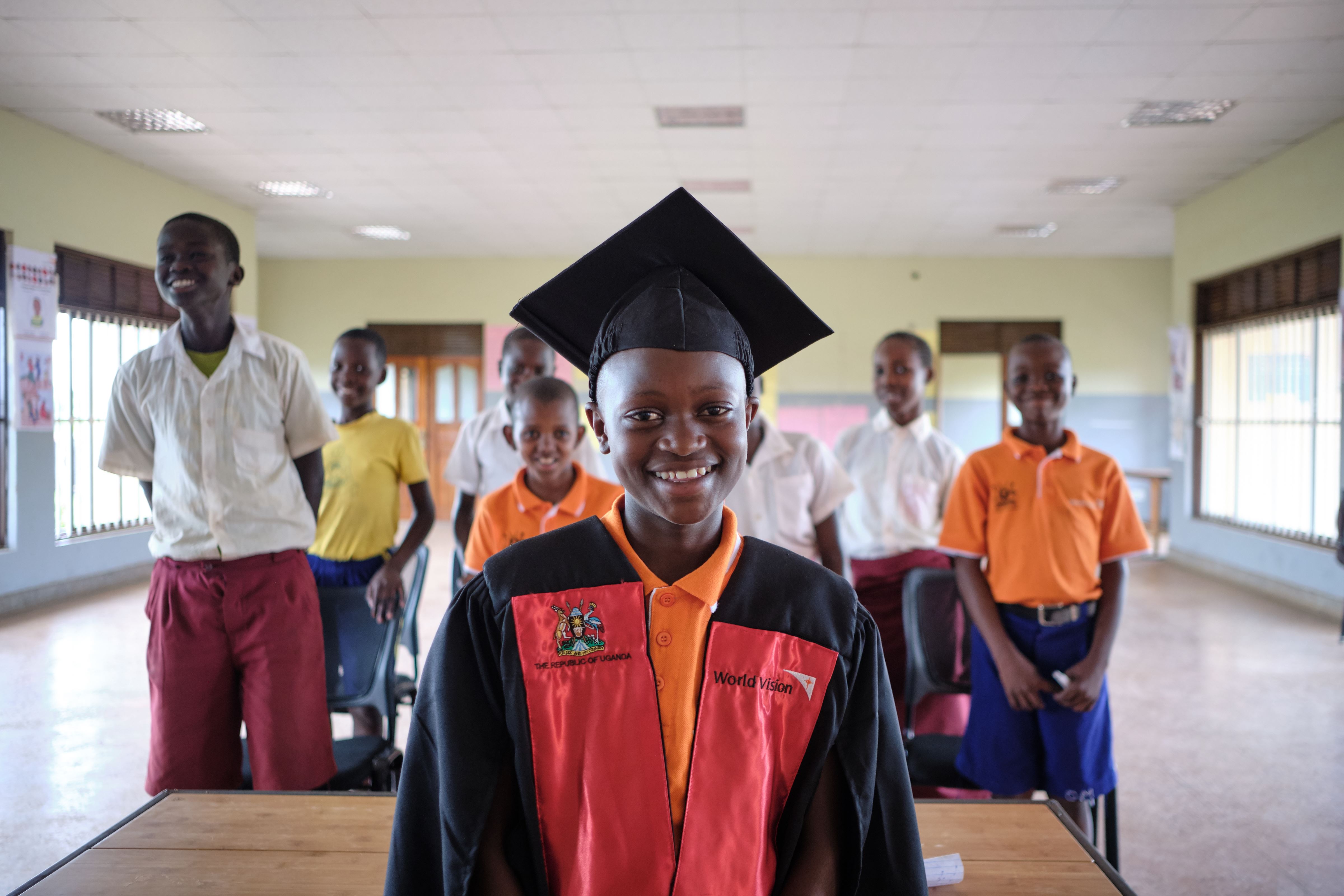 Un niño ugandés con un gorro de graduación en la parte delantera de una clase, detrás de él se ve a otros estudiantes.