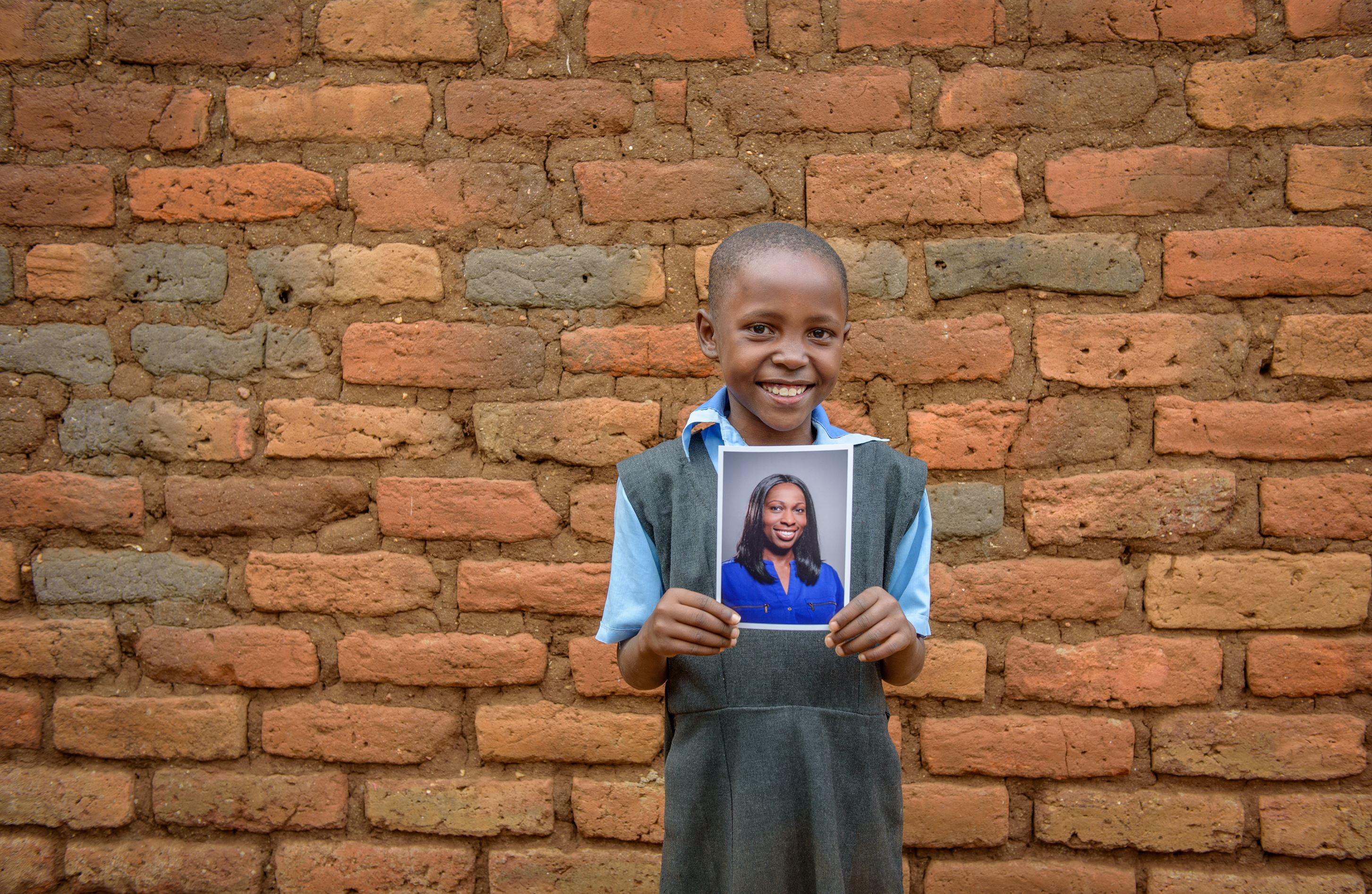 Niña de Kenia de pie frente a una pared de ladrillos, sosteniendo la foto de su padrino y sonriendo.