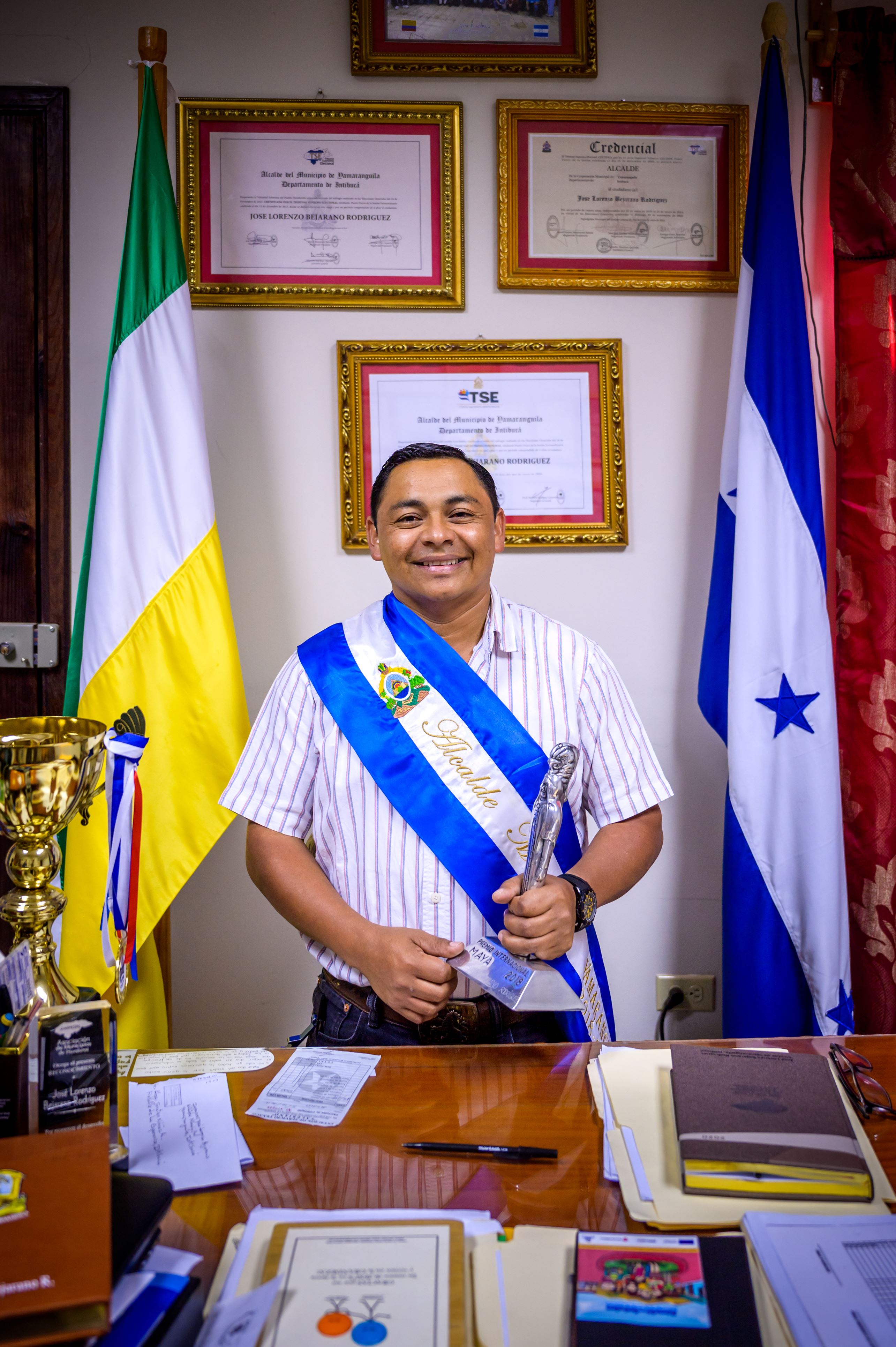 Hombre de Honduras que lleva una banda y sostiene un trofeo mientras está de pie en su oficina entre dos banderas