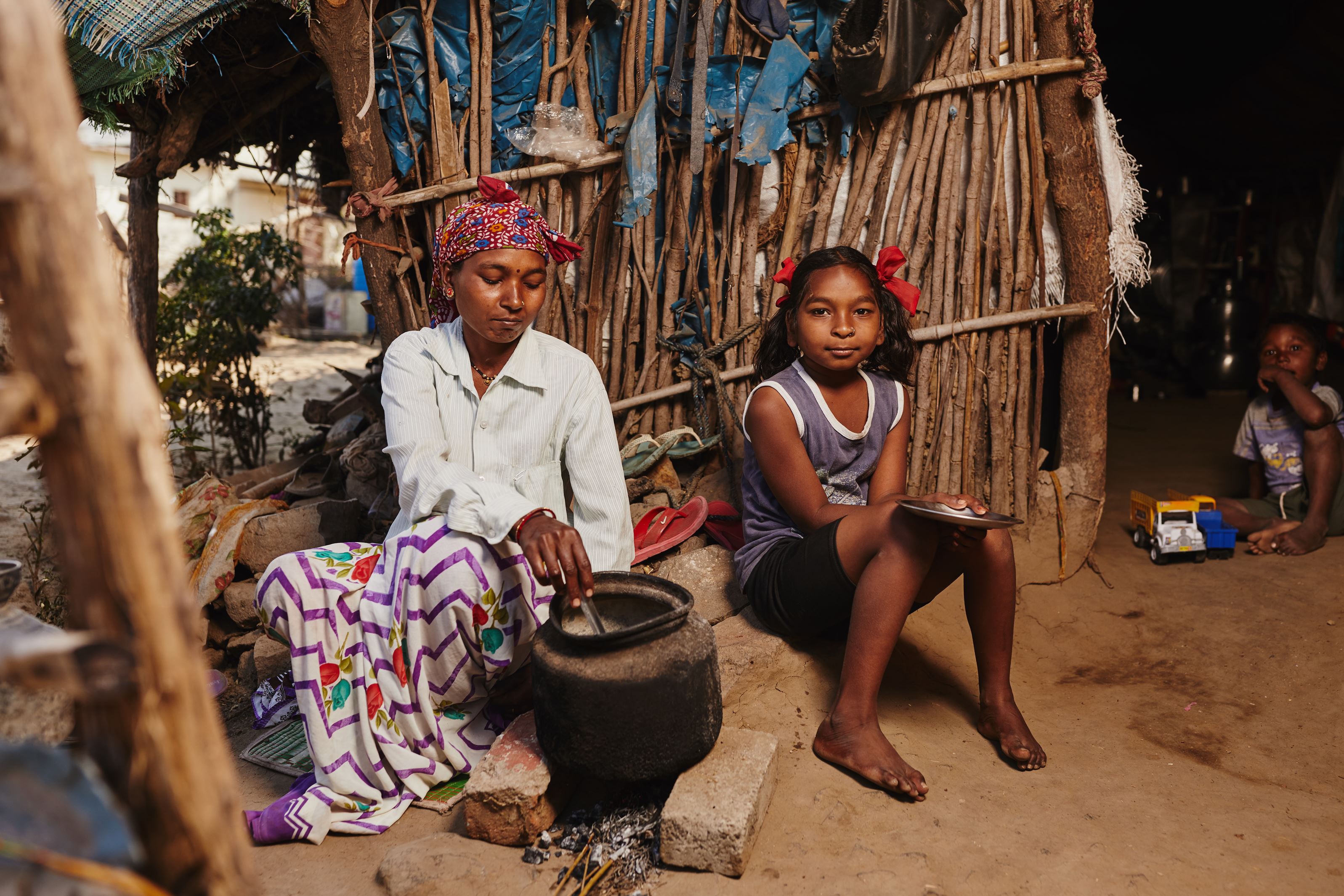 Niña de la India, descalza y con su madre sentada fuera de su casa mientras su madre cocina.