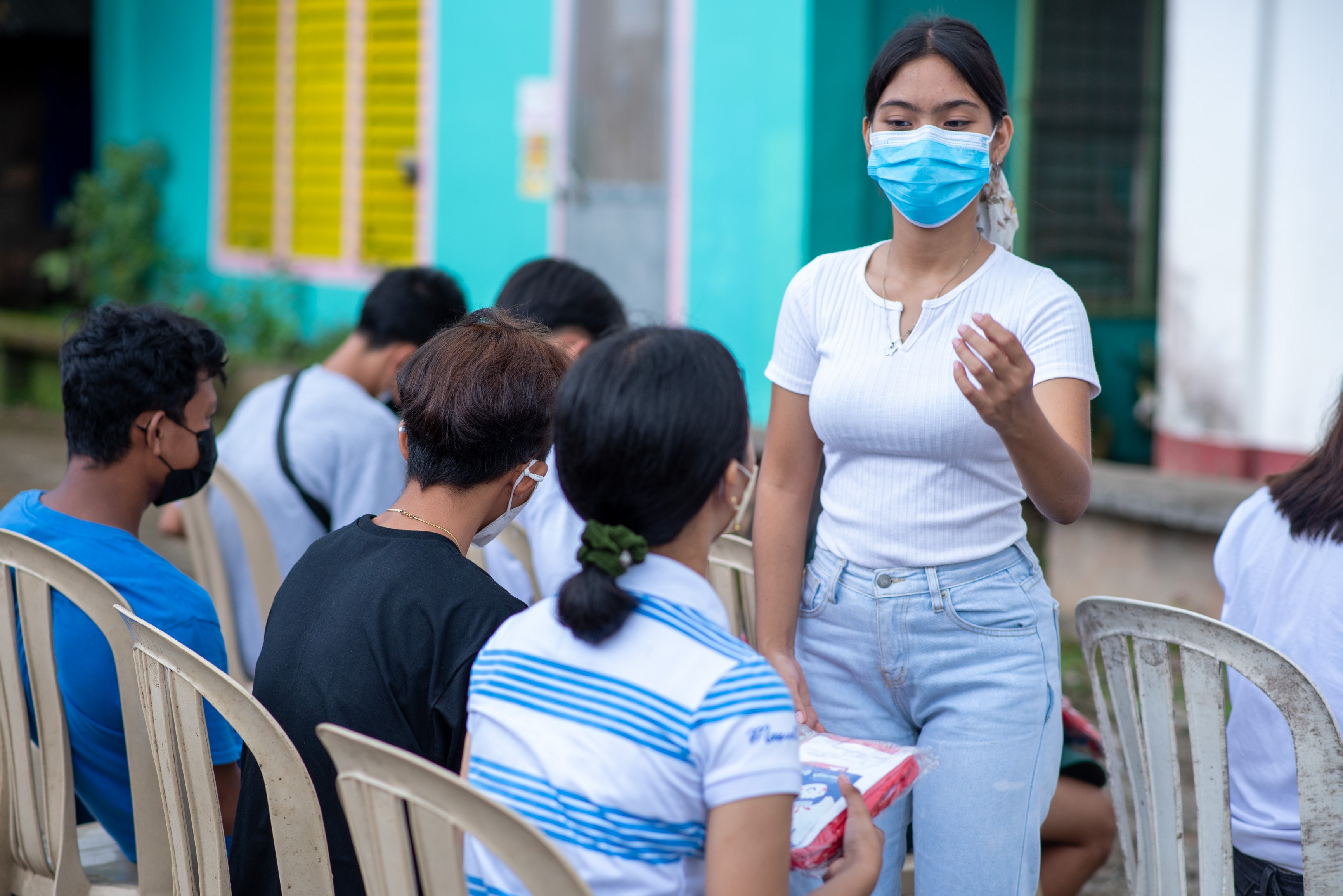 Una adolescente filipina con una mascarilla quirúrgica habla a un grupo de personas sentadas en sillas de plástico, de espaldas a la cámara.