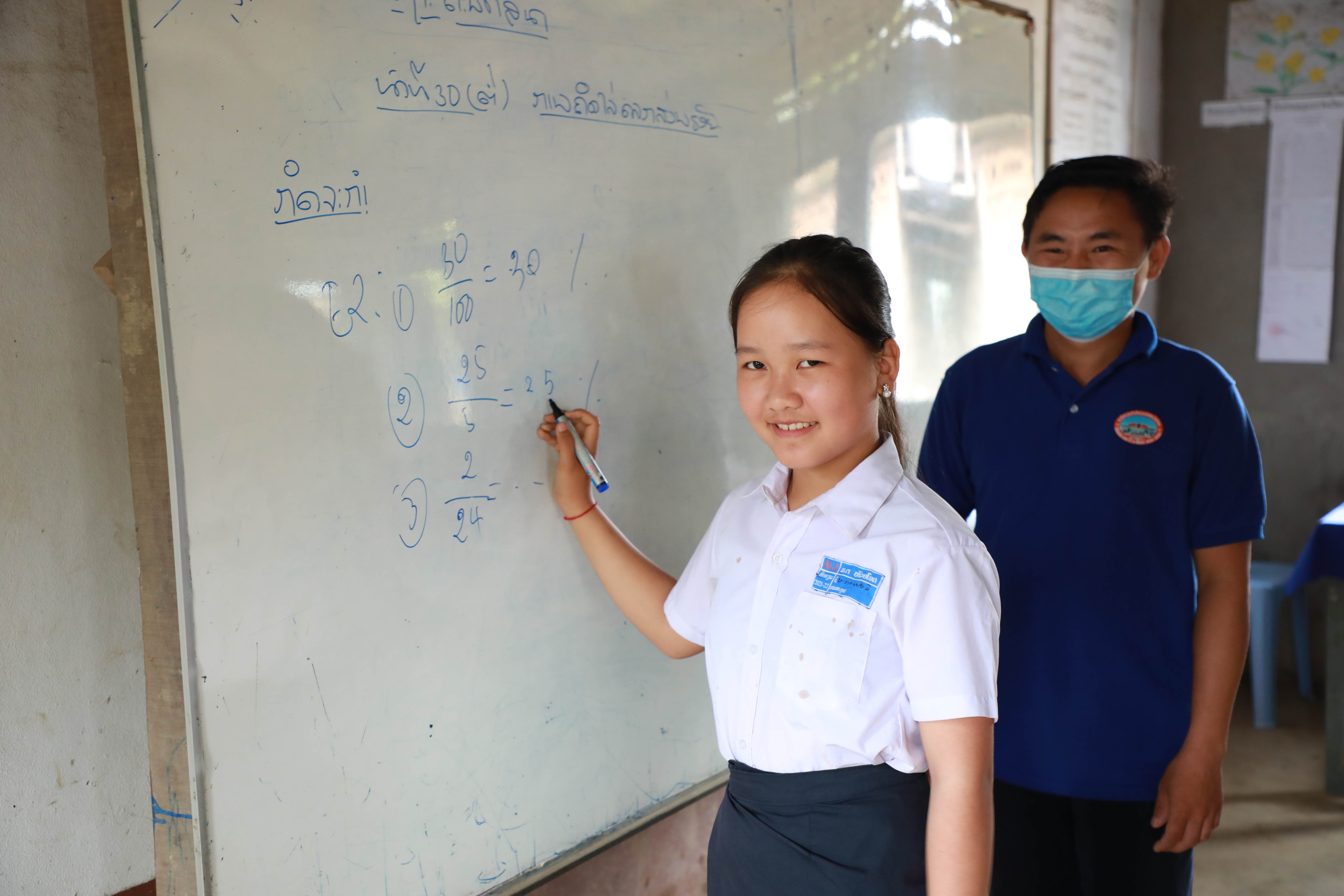 Una estudiante de Laos escribe en una pizarra en la escuela mientras sonríe a la cámara. Su profesor supervisa en el fondo mientras lleva una mascarilla.