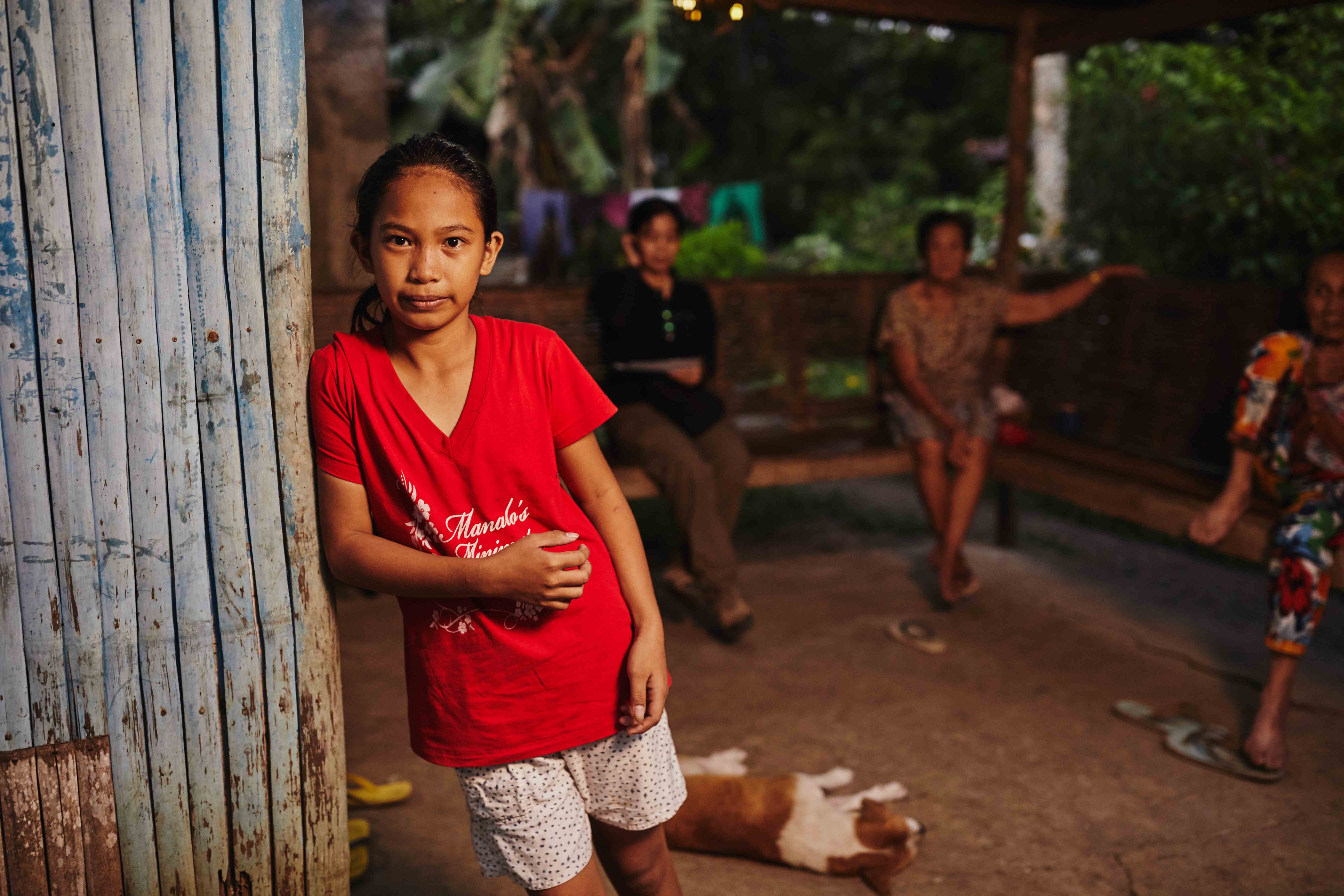 Niña filipina apoyada en un panel metálico mientras mira a la cámara y su familia es visible en el fondo