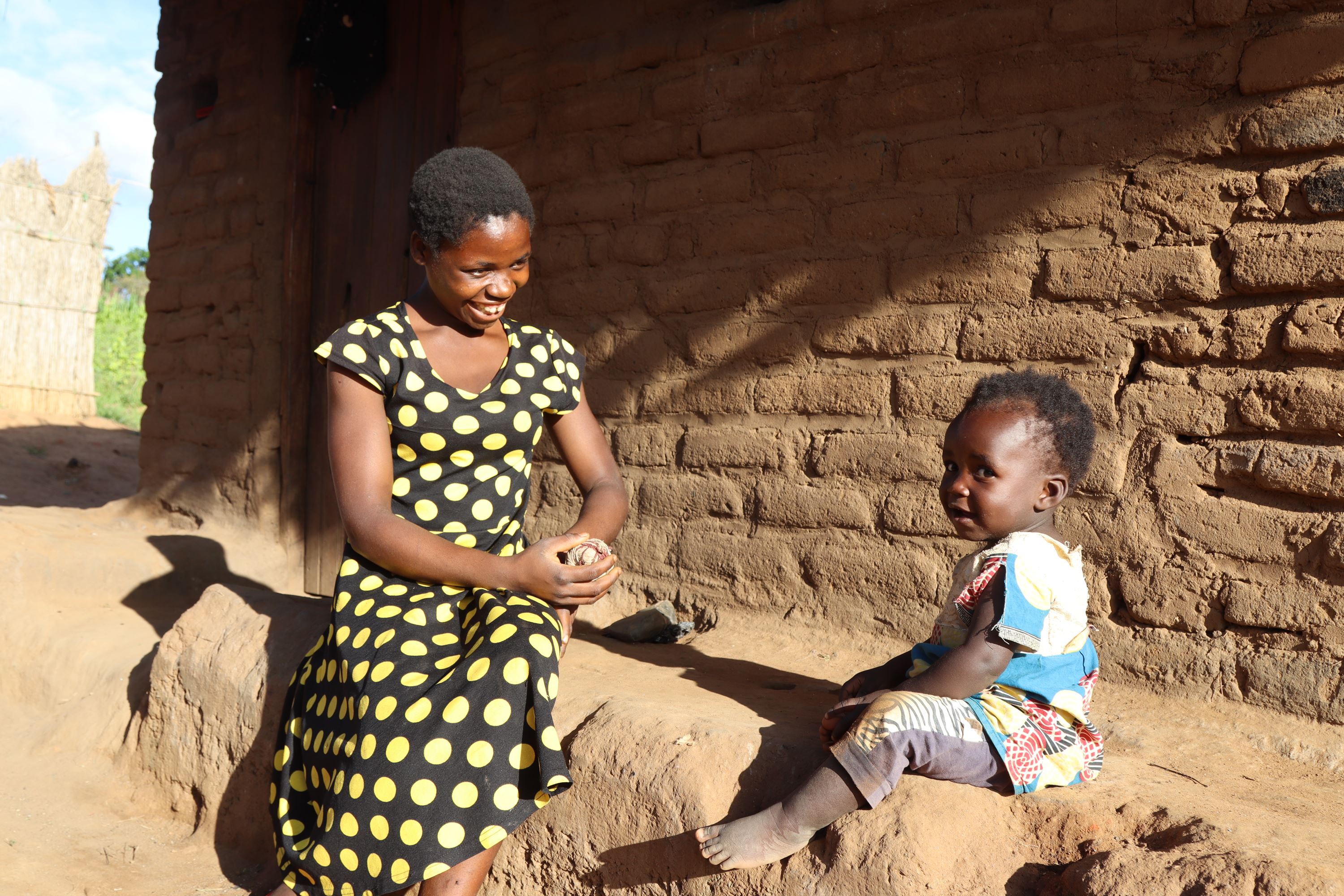 Grace, una madre adolescente de Malawi, con un vestido de lunares negros y amarillos, se sienta junto a su hija pequeña y le sonríe.